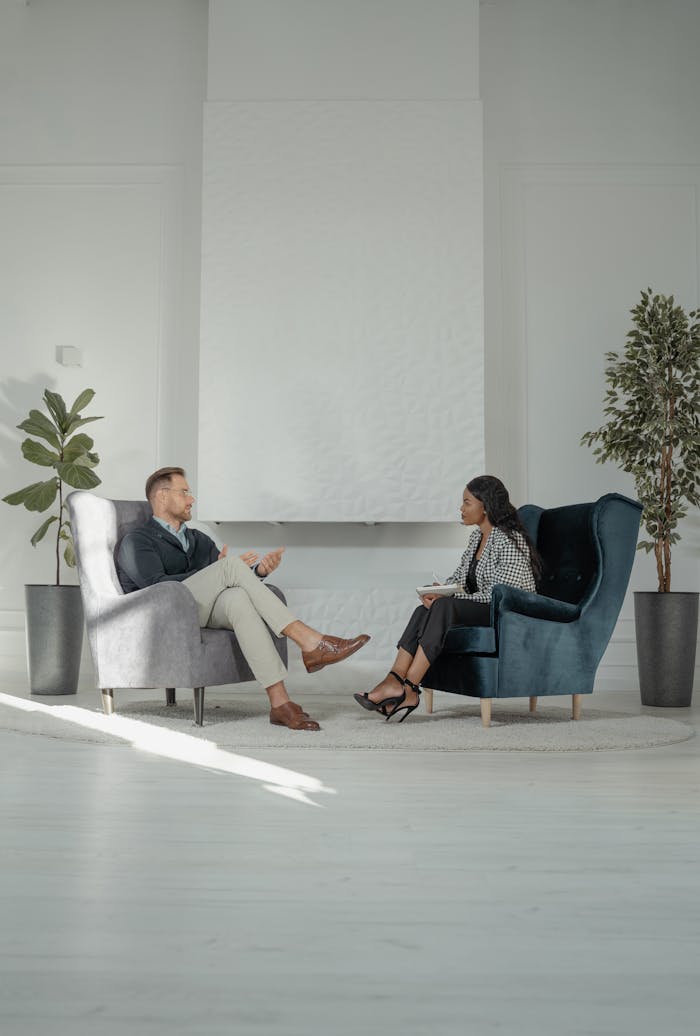 Two professionals in a conversation, sitting on armchairs with a white background, indoors.